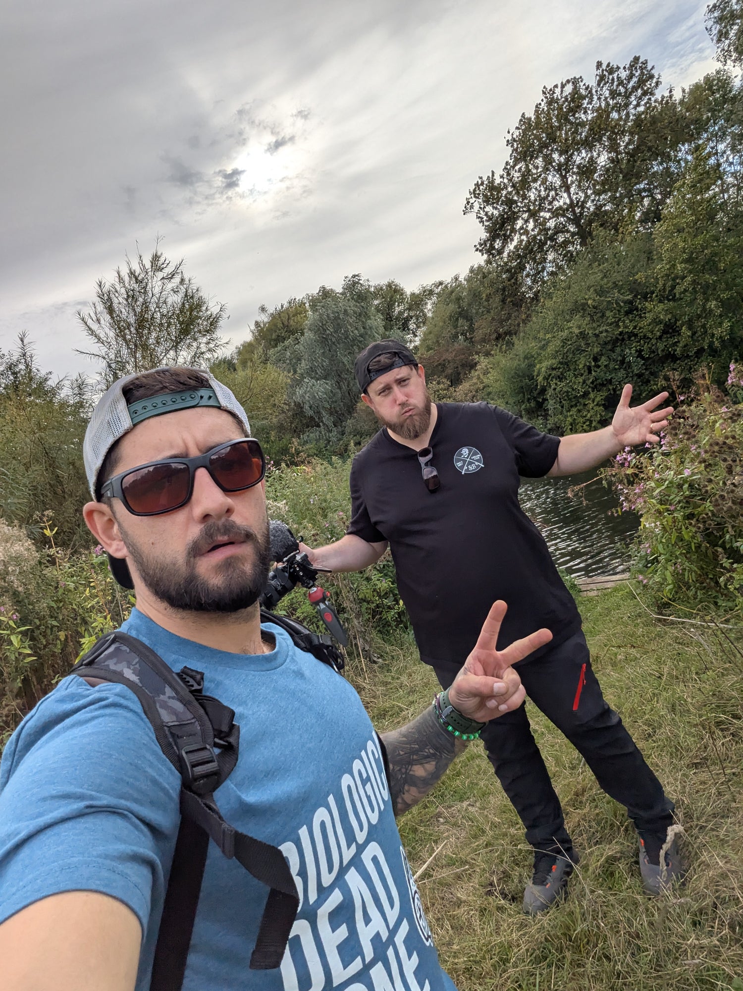 Two men by a river in nature holding filming and fishing equipment