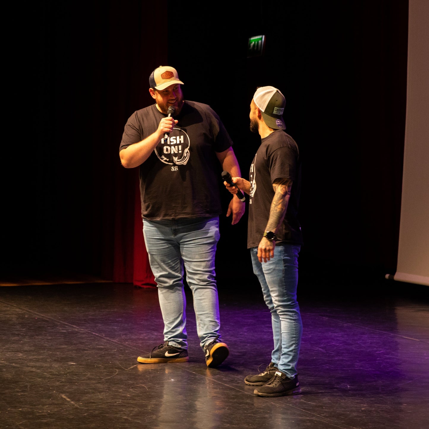 Two men on stage with one holding a microphone, in front of a dark background with a screen displaying text.