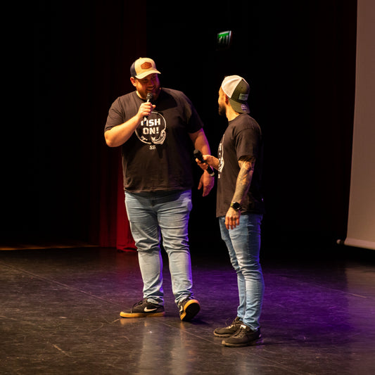 Two men on stage with one holding a microphone, in front of a dark background with a screen displaying text.