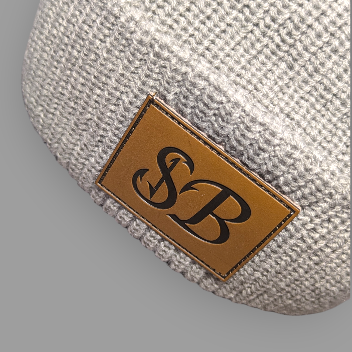 Textured beige fabric pocket square with a brown leather patch featuring 'SB' initials on a gray background.
