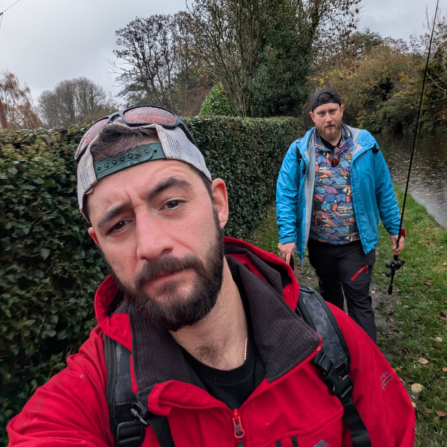 Two men outdoors, one taking a selfie, with a hedge and trees in the background. going fishing.